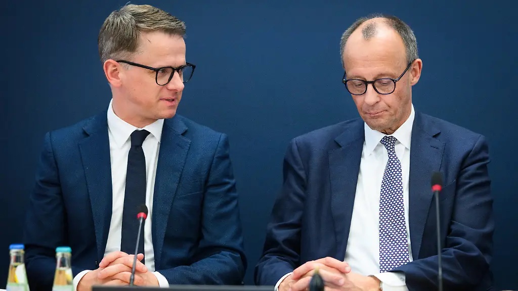 Carsten Linnemann and Friedrich Merz seated side by side at a table with microphones against a dark-blue background