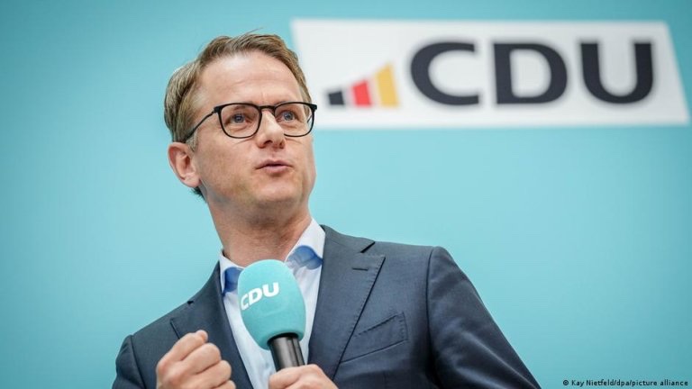 Carsten Linnemann speaking into a CDU microphone in front of a CDU logo on a turquoise backdrop