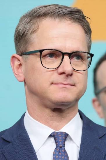 Close portrait of Carsten Linnemann in dark-rimmed glasses and a navy suit against a turquoise and orange background