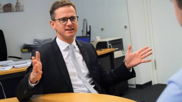 Carsten Linnemann seated at a desk in an office, gesturing during a conversation, with files and a printer in the background