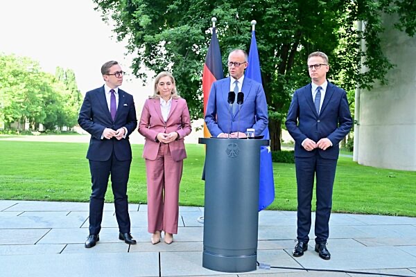 Chancellor Friedrich Merz speaking at an outdoor lectern, with Carsten Linnemann standing to the right and two other people to the left, German and EU flags behind