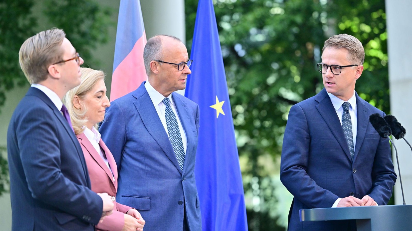 Carsten Linnemann speaking at an outdoor lectern, with Friedrich Merz and two other people standing to the left, German and EU flags behind