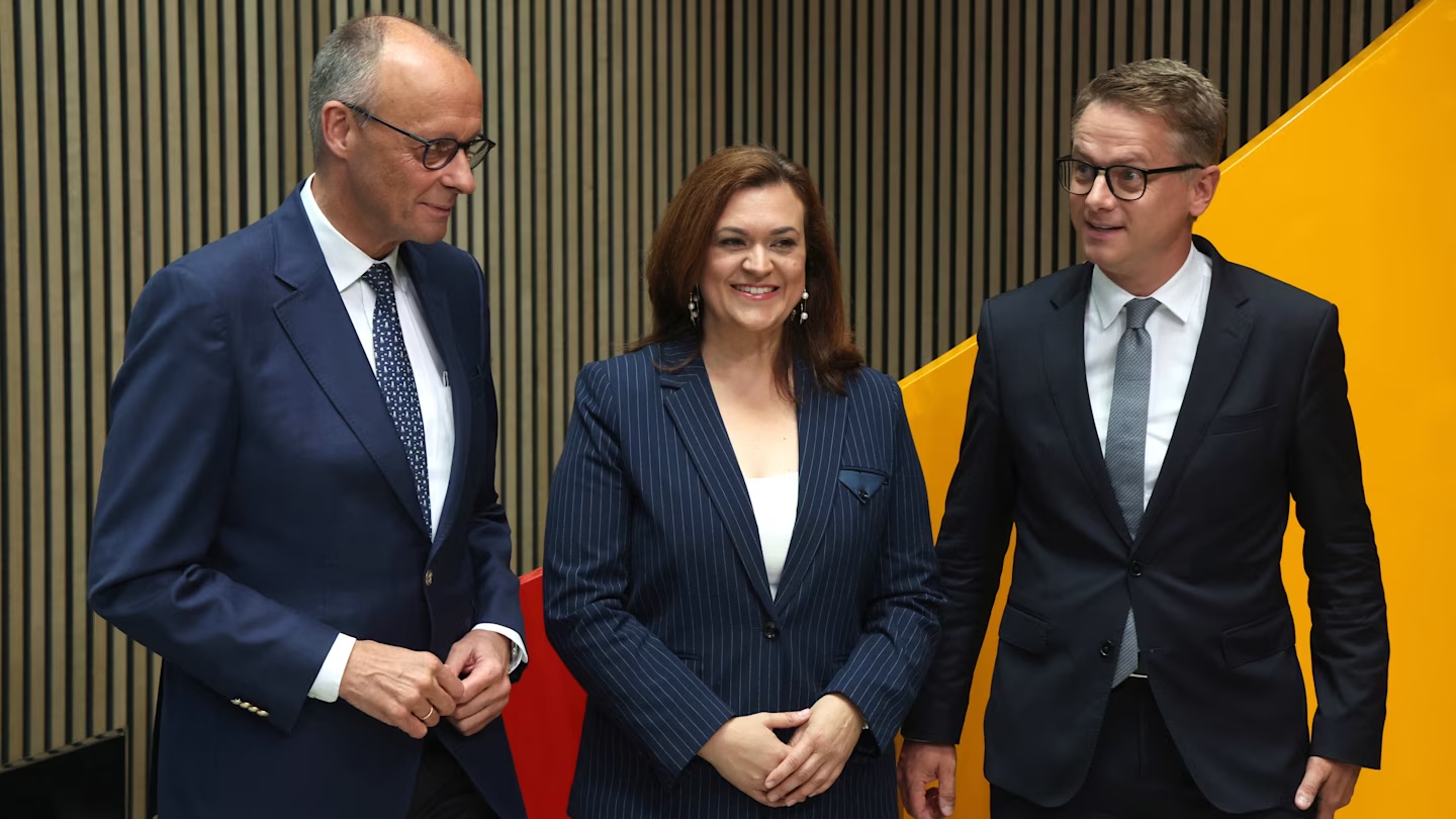 Friedrich Merz, a woman and Carsten Linnemann standing side by side in front of a wooden wall and a yellow panel