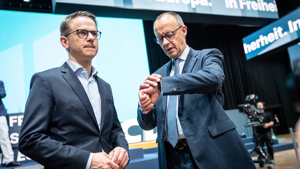 Carsten Linnemann and Friedrich Merz at a CDU event; Merz checks his watch in front of a backdrop reading “Europa. In Freiheit”